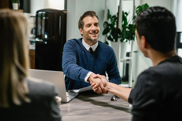 A hiring manager in a blue sweater enthusiastically shakes hands with a candidate in an office setting.