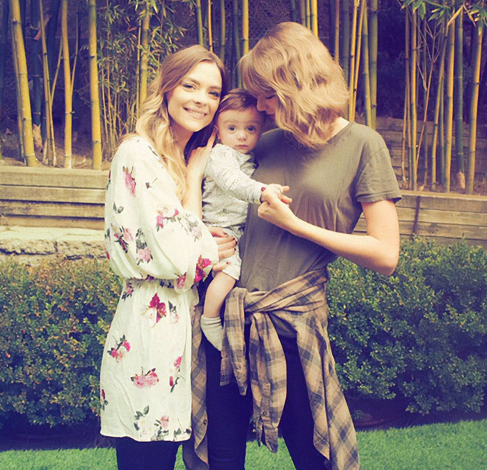 Two women outdoors holding a baby, one in floral, the other in a green shirt, representing Taylor Swift's godson situation.