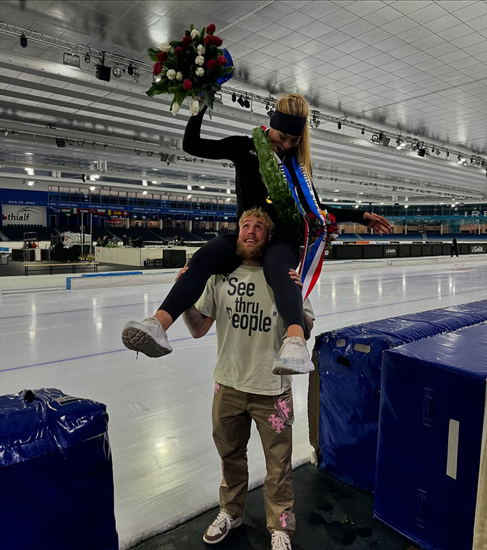 Man in arena lifting a speed skater with flowers, wearing a "See thru People" shirt.