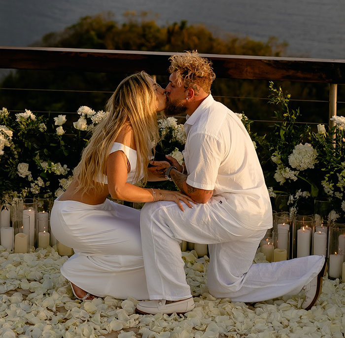 Couple embraces in a romantic proposal setting decorated with white flowers and candles, featuring Jake Paul.
