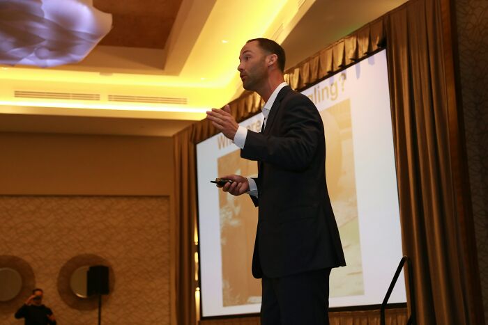 Man in a suit speaking at a wedding-related event, with a presentation behind him.