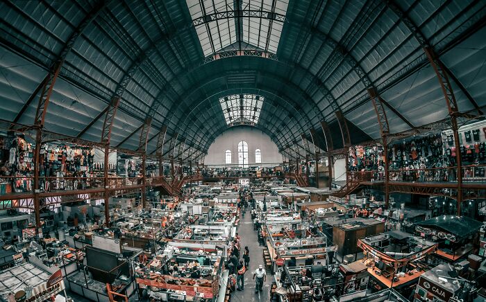 Busy indoor market with high arched ceilings, showcasing bustling people and various booths and stalls.