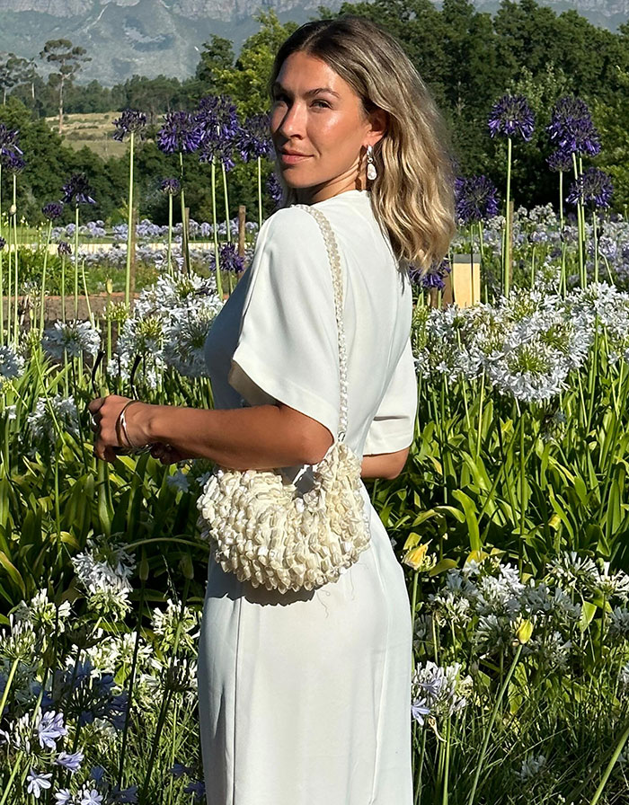 Woman in elegant white dress standing in a garden, holding a textured bag, with purple flowers in the background.