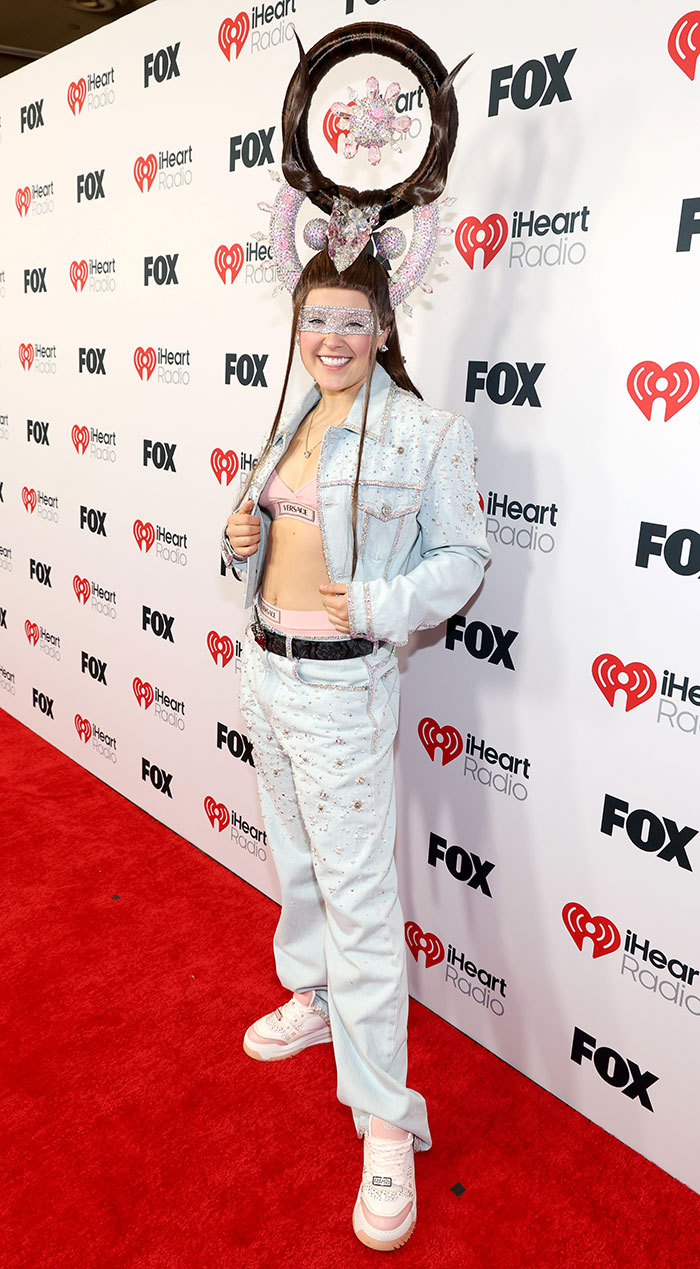 Person on 2025 iHeartRadio red carpet in a wild outfit with a large, intricate headpiece and a bedazzled denim ensemble.
