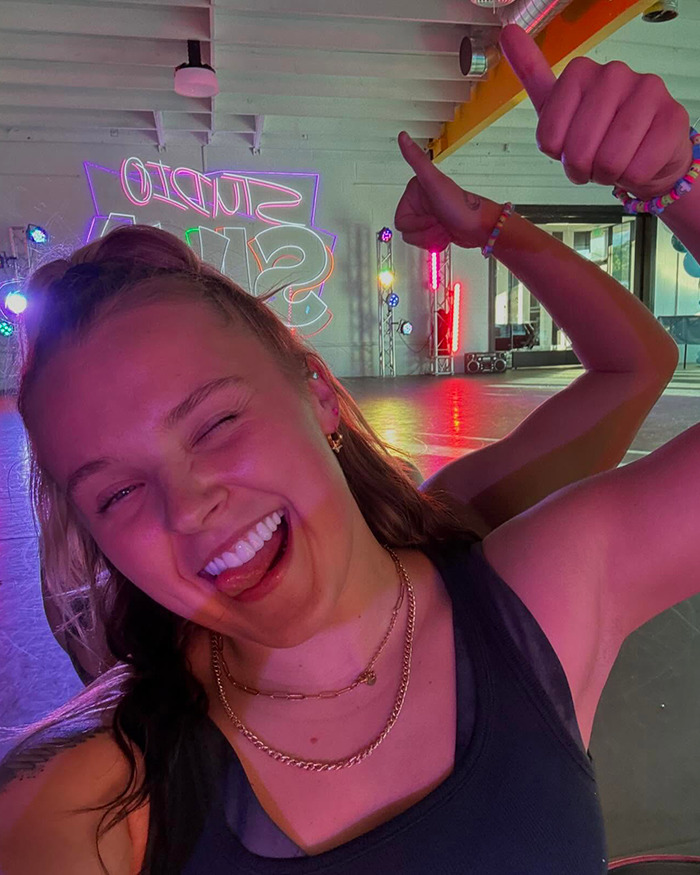 Person inside dance studio smiling with thumbs up, vibrant lights in the background, showcasing new style.