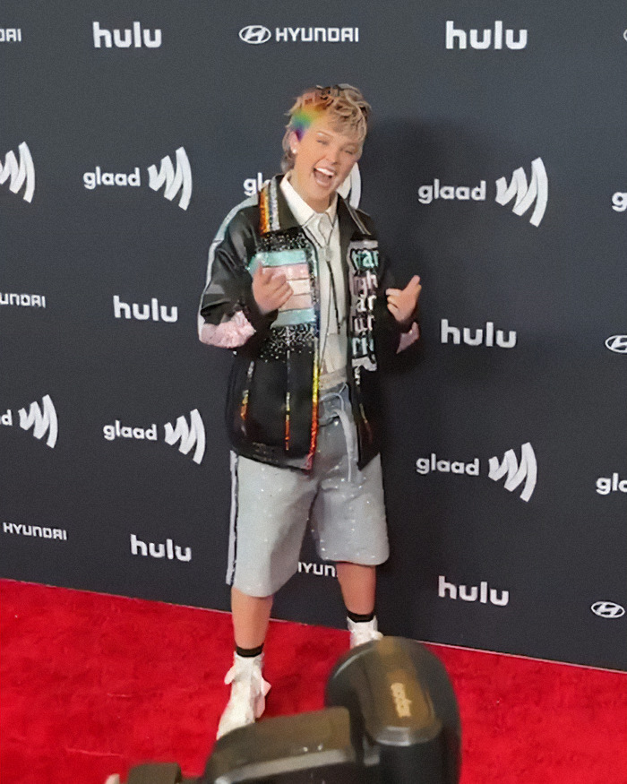 JoJo Siwa debuts a shaved rainbow head at a GLAAD event, wearing a colorful jacket and shorts on the red carpet.