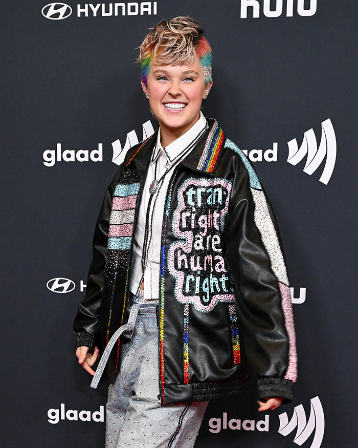 JoJo Siwa with a shaved rainbow head, wearing a colorful jacket promoting LGBTQ+ rights, at a public event.