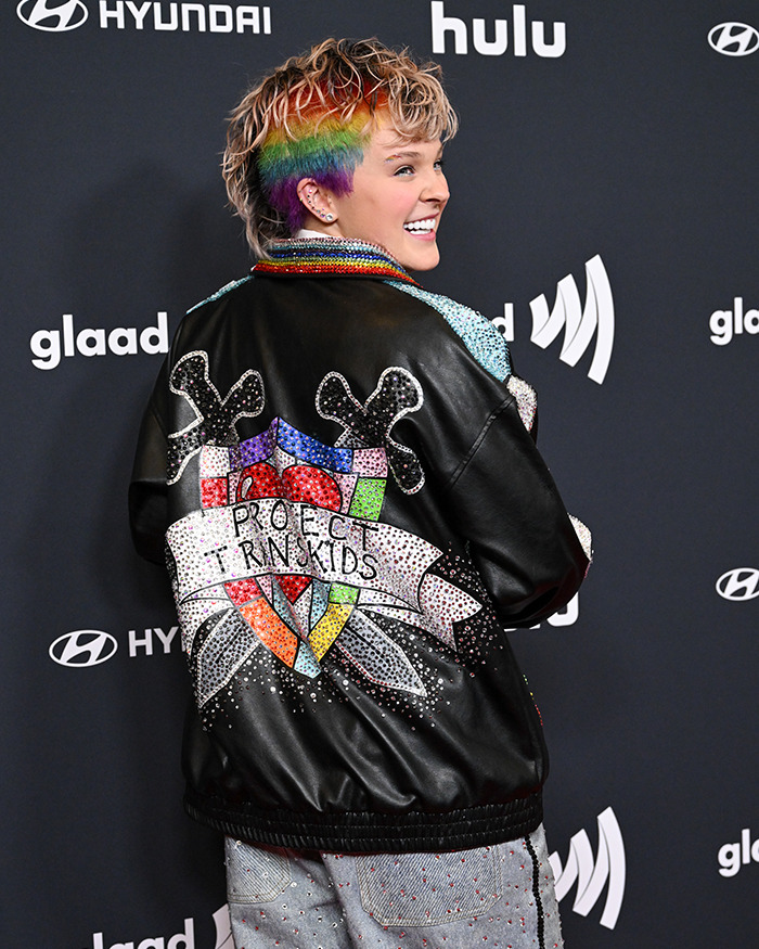 JoJo Siwa in a rainbow shaved hairstyle shown from the back, wearing a decorative jacket at a public event.
