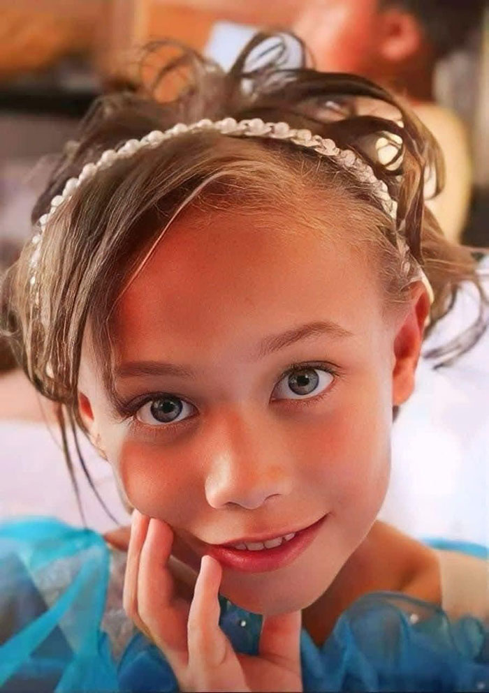 Young girl with light eyes and skin, wearing a blue dress and a headband, smiling at the camera.