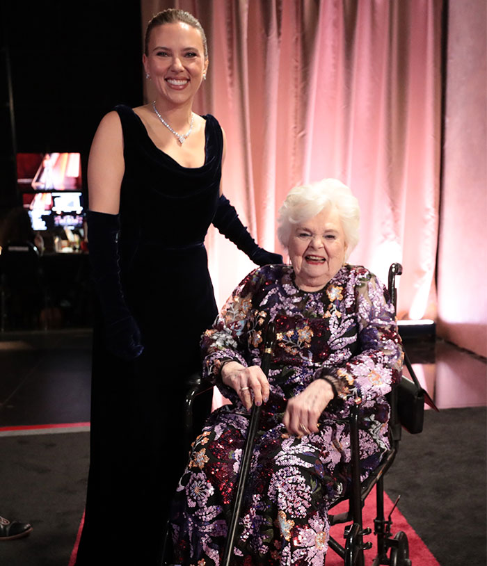 Two women backstage at Oscars 2025, one in a black dress, the other seated in a wheelchair wearing a floral gown.