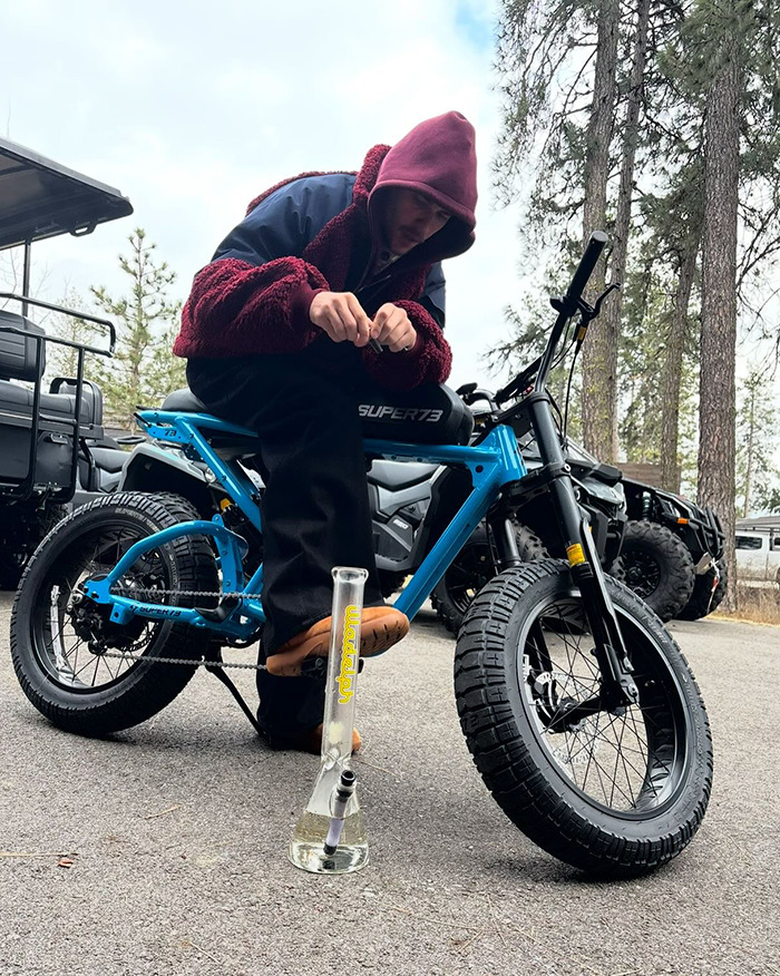 Justin Bieber in a hood sitting on a blue bicycle outdoors.