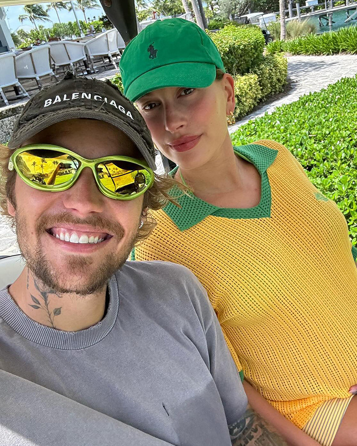 Hailey Bieber and Justin Bieber taking a selfie outdoors, one wearing a yellow shirt and green cap, the other in a gray shirt and sunglasses.