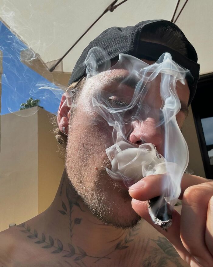 A person with tattoos and a cap smoking, with visible smoke swirling around, outdoors under a clear sky.
