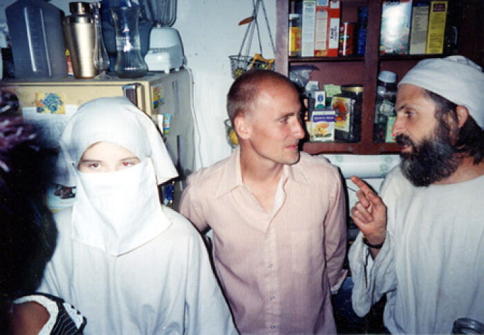 Three people in a kitchen, one in white covering, discussing next to shelves. Creepy photo with a dark history vibe.