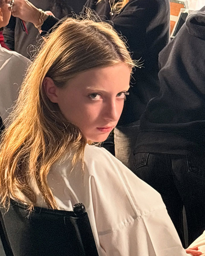 Young model at Paris Fashion Week, sitting in a chair, wearing a white outfit, hair styled, surrounded by people in the backstage.