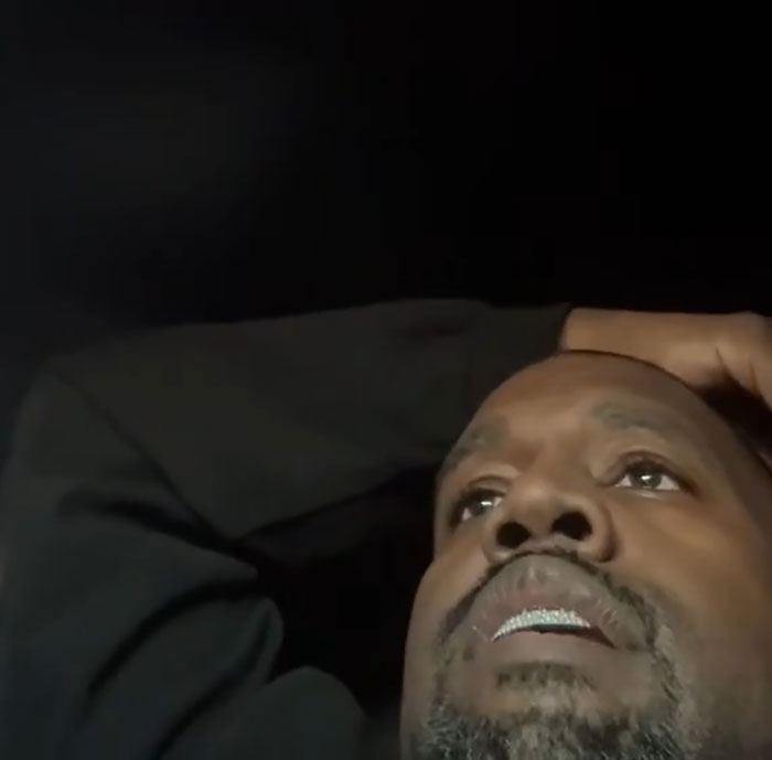 Man lying down, wearing a black shirt, looking thoughtful against a dark background.