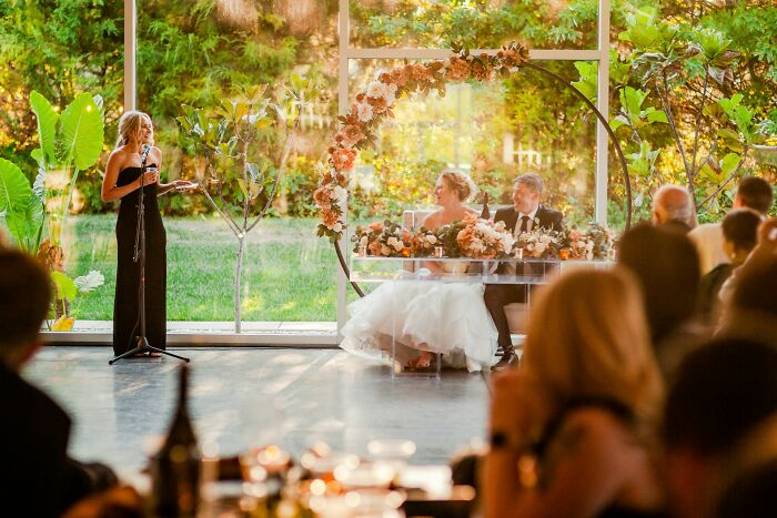 Wedding scene with a guest speaking, bride and groom at a table, surrounded by floral decorations and natural light.