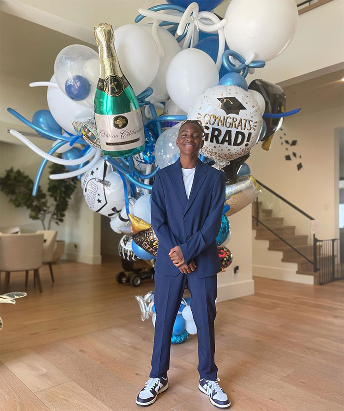 Kevin Hart in blue suit stands in front of graduation balloons at a party, smiling. Kevin Hart in blue suit stands in front of graduation balloons at a party, smiling.