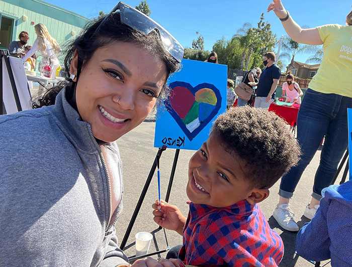Eniko Hart smiling with a child at an outdoor art event, showcasing a colorful heart painting. Eniko Hart smiling with a child at an outdoor art event, showcasing a colorful heart painting.