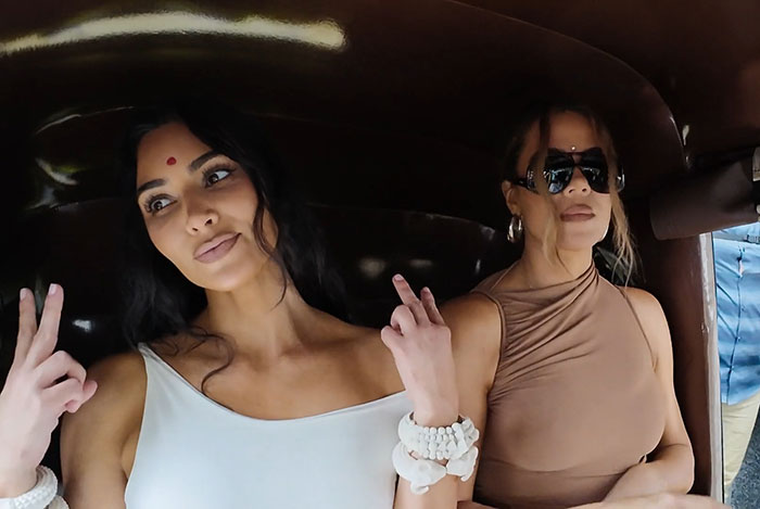 Two women in a vehicle, one with a bindi, posing and gesturing, reflecting modern cultural fashion.