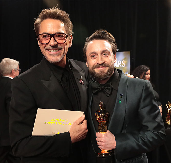 Two men smiling backstage at Oscars 2025, one holding an envelope and another with a trophy, both in formal wear.
