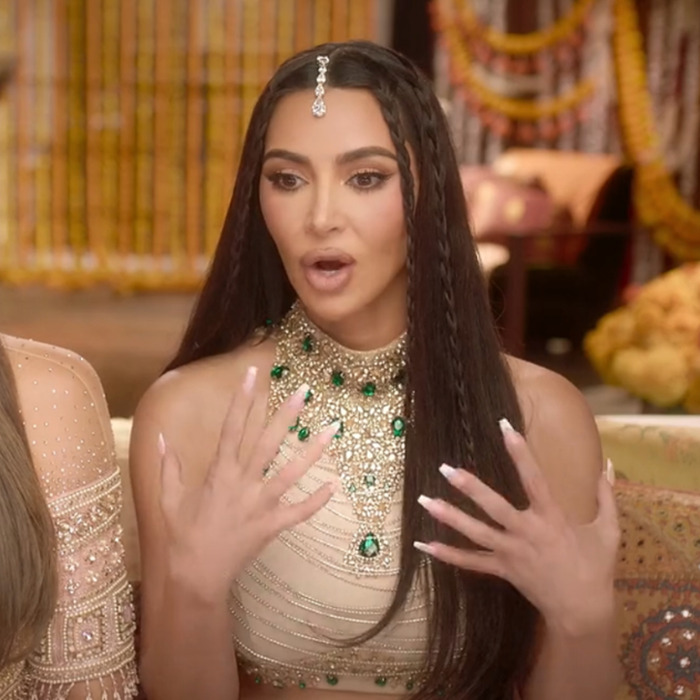 Woman expressing surprise, wearing a jeweled top and headpiece, hands raised with painted nails.