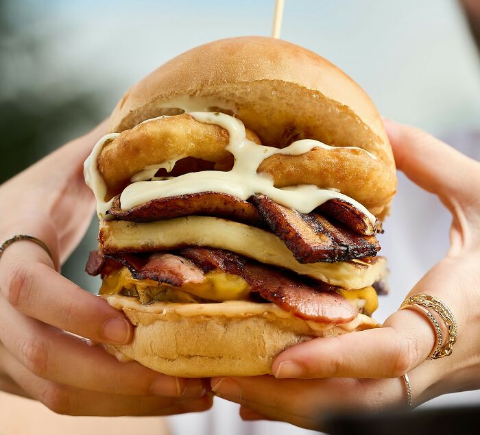Burger with bacon, cheese, and onion rings in hands, a controversial meal some find disgusting but enjoyed worldwide.