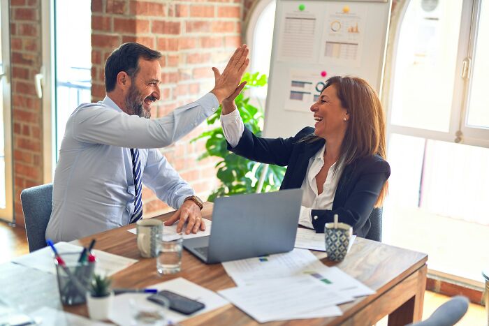 Two colleagues high-fiving in an office, surrounded by documents and a laptop, expressing unity in work achievements.
