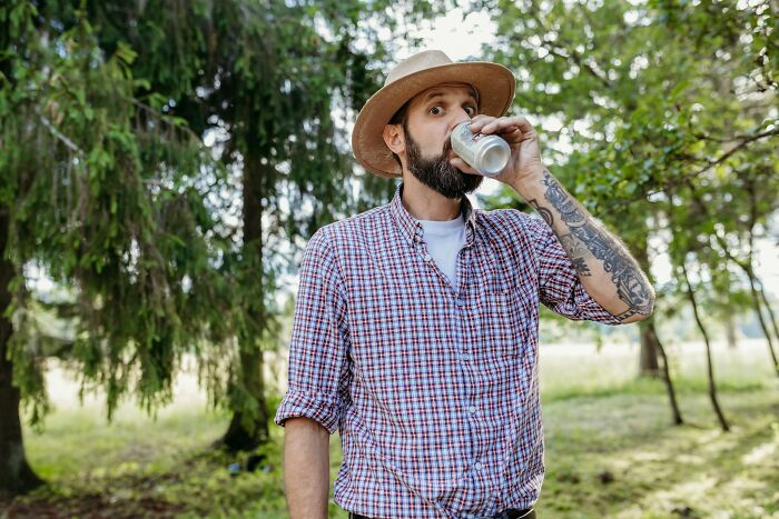Man in a hat and checkered shirt drinking from a can in a forest, showcasing casual style.