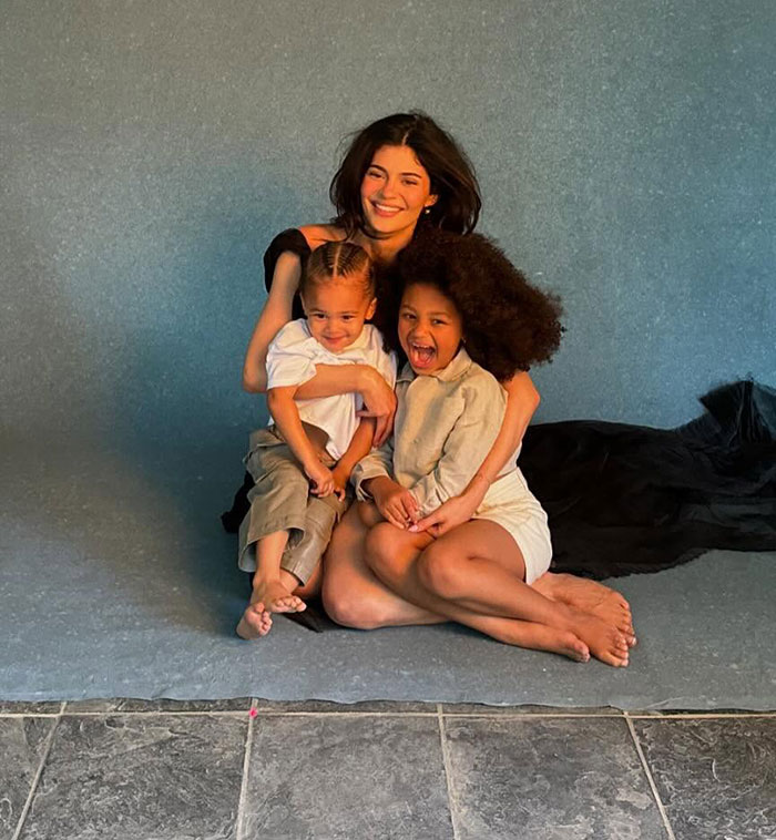 Woman smiling with two children against a plain backdrop, seated on the floor, representing family warmth and connection.