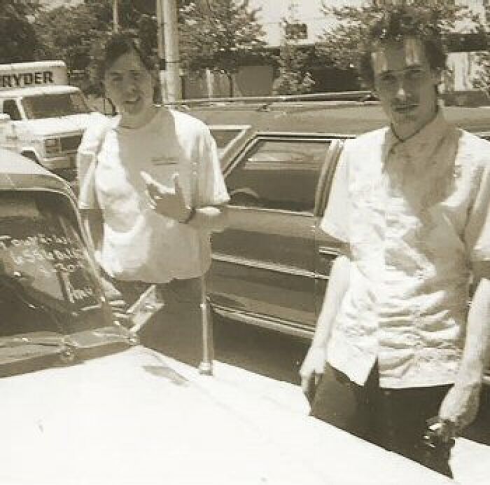 Two people in a vintage photo stand near a car in a parking lot, capturing a moment with a creepy history.