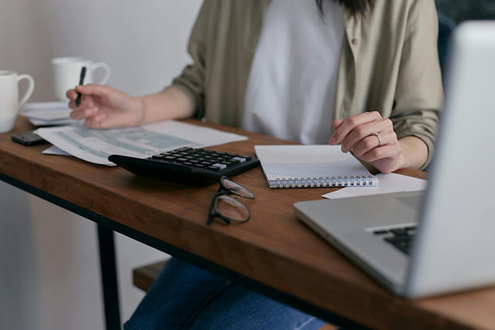 A woman at a desk using a calculator and notebook, with a laptop and glasses nearby, handling financial matters.