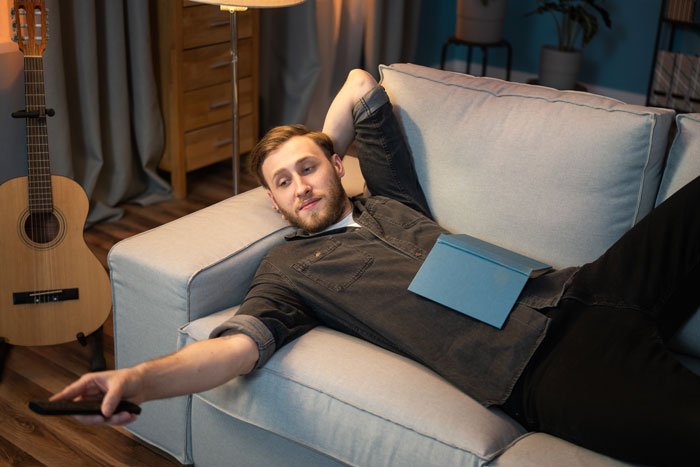 Man relaxing on a couch, symbolizing laziness, with a closed book on his chest and a remote in hand.