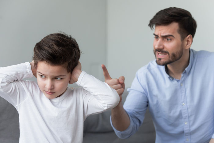 Man lecturing a boy about laziness while the boy covers his ears at home.