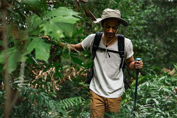 Man hiking in a dense forest, wearing a hat and carrying a trekking pole, demonstrating life-saving outdoor skills.