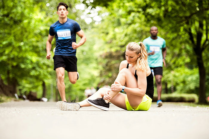 Runner sitting on ground, holding her injured knee, while two other runners approach on a tree-lined path.