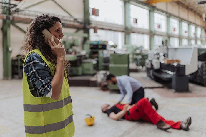 Woman in a safety vest on phone, emergency aid scene with injured worker on factory floor.