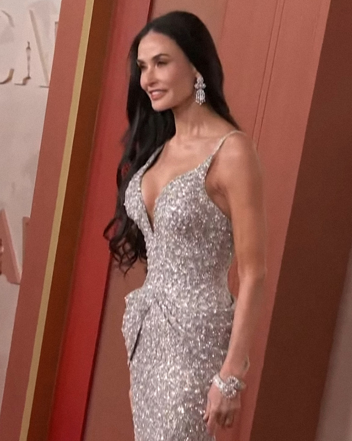 Demi Moore in a glittering silver gown at an award event, with long brunette hair and statement earrings.