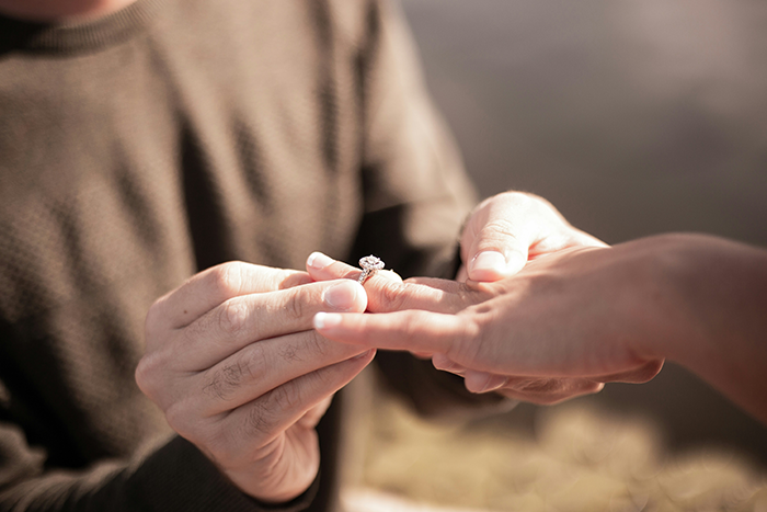Hands exchanging a diamond ring outdoors, symbolizing an engagement or wedding commitment.