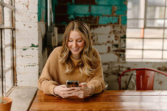 Woman smiling at her phone in a cozy cafe setting, representing a moment of relief after leaving a friend's chaotic wedding plans.