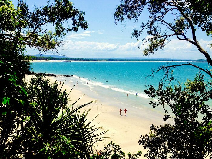 Stunning beach view with white sand and clear turquoise waters, framed by lush green foliage and distant horizon.