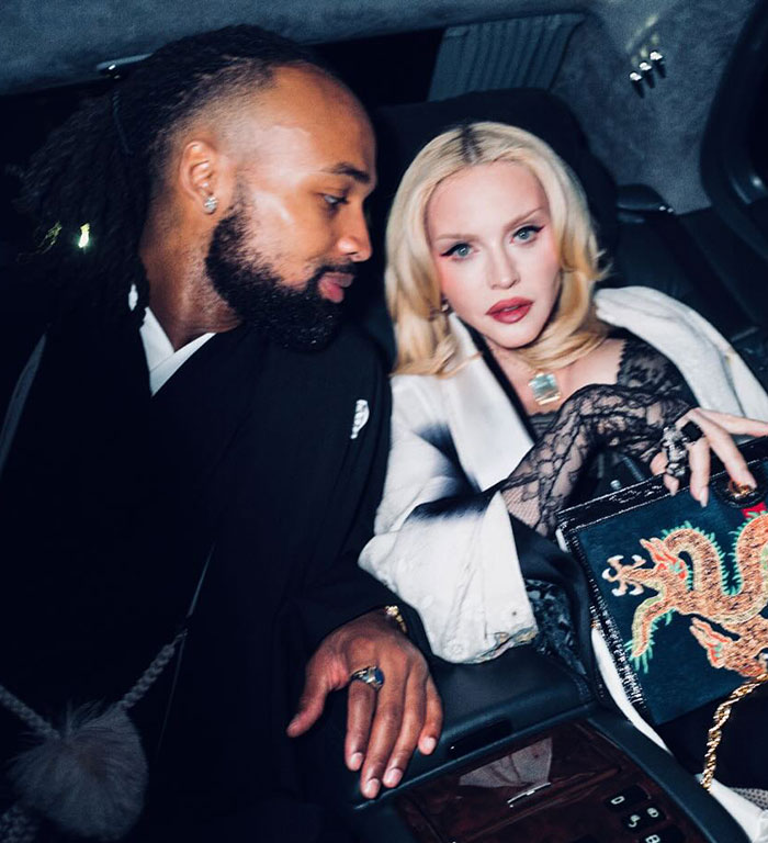 Blonde woman with smooth face holds embellished purse while sitting in a vehicle with a man.