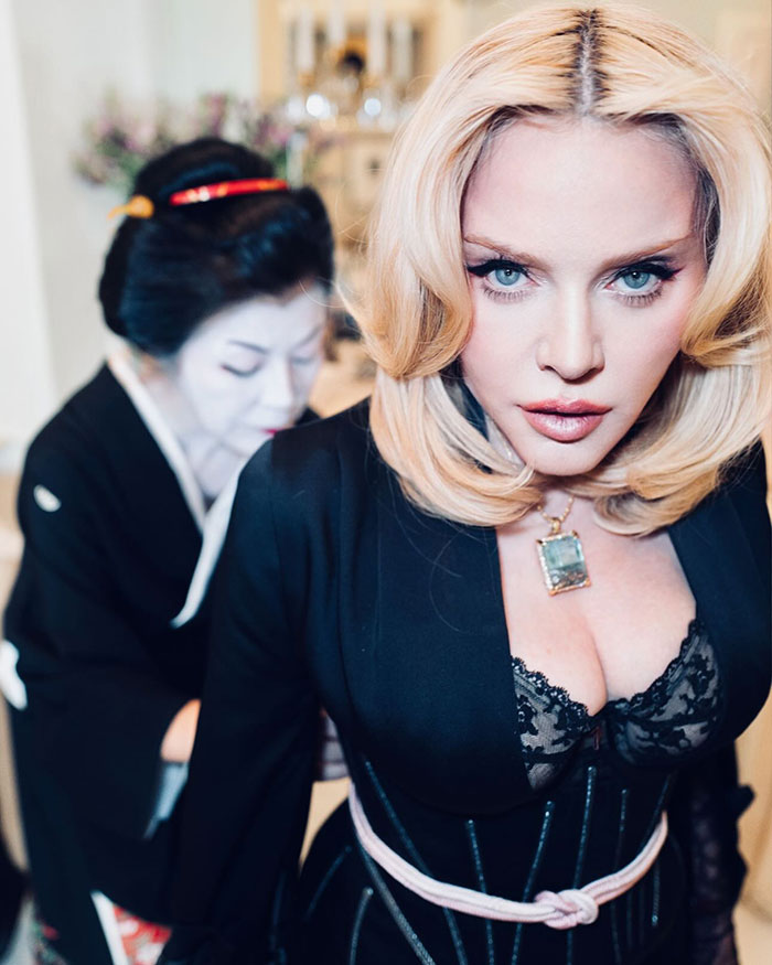 Madonna with smooth face, wearing a black outfit and necklace, in a room with another person in the background.