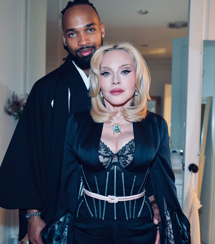 Madonna with smooth face and an individual in a black outfit, standing together in a softly lit room.