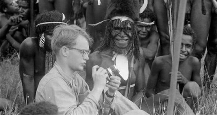 Man examining a camera surrounded by indigenous people, hinting at a seemingly normal yet terrifying backstory.