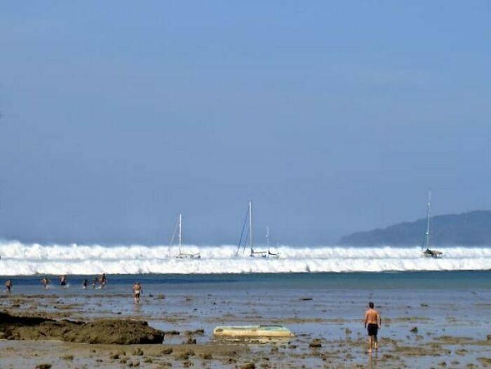 Playa con personas y barcos, escena normal que oculta una historia inquietante de un tsunami acercándose.