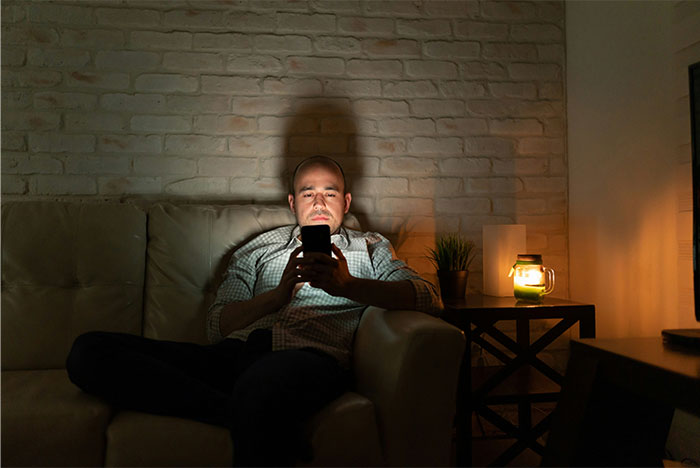 Man using smartphone on sofa, dimly lit room, discussing male loneliness epidemic solution online.