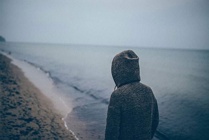 Person in a hooded sweater stands alone by the sea, reflecting on the male loneliness epidemic.