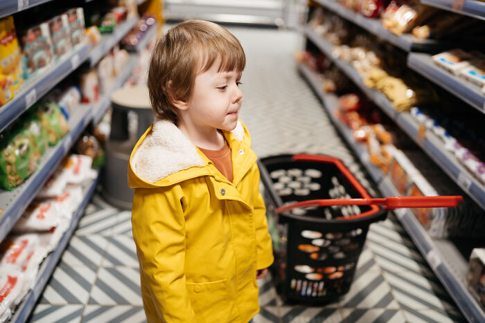 Young child in yellow coat in supermarket aisle, standing beside a shopping basket filled with items. Young child in yellow coat in supermarket aisle, standing beside a shopping basket filled with items.