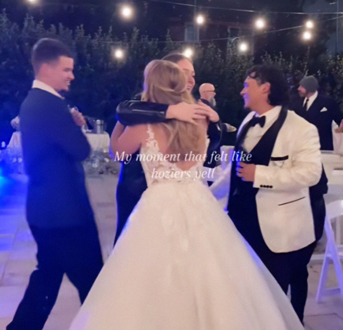 Couple embraces at wedding reception, capturing guests' surprise proposal reactions under string lights.